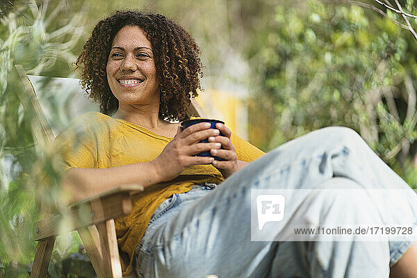 Lächelnde  lockig behaarte Frau mit Kaffeetasse  die wegschaut  während sie auf einem Stuhl im Gemüsegarten sitzt