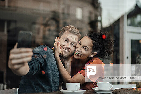 Freund nimmt Selfie mit Freundin durch Handy beim Sitzen im Cafe