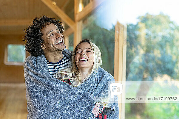 Glückliches Paar  das in eine Decke gehüllt ist und lächelnd am Fenster im Vorgarten steht