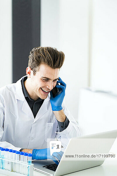 Lächelnder Wissenschaftler  der mit seinem Smartphone spricht  während er einen Laptop im Labor benutzt