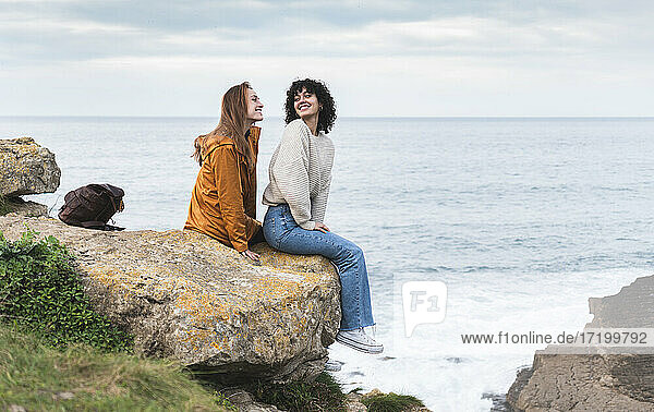 Lächelnde Freundinnen  die sich gegenseitig ansehen  während sie auf einer Klippe am Meer sitzen