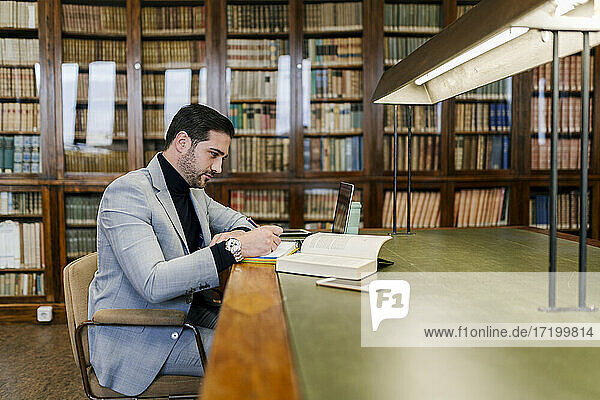 Geschäftsmann mit Laptop  der in einem Buch schreibt  während er in einer Bibliothek sitzt
