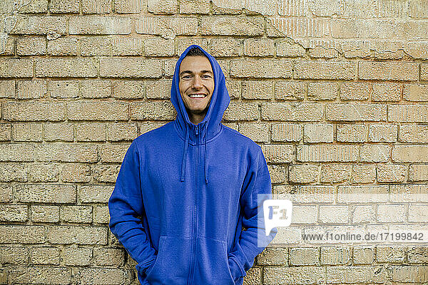 Lächelnder männlicher Sportler in blauer Jacke vor einer Backsteinmauer