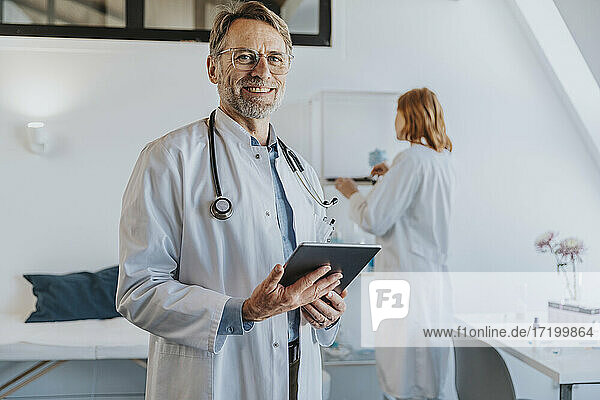 Lächelnder Arzt mit digitalem Tablet  der mit einem Mitarbeiter im Hintergrund in einer Klinik steht