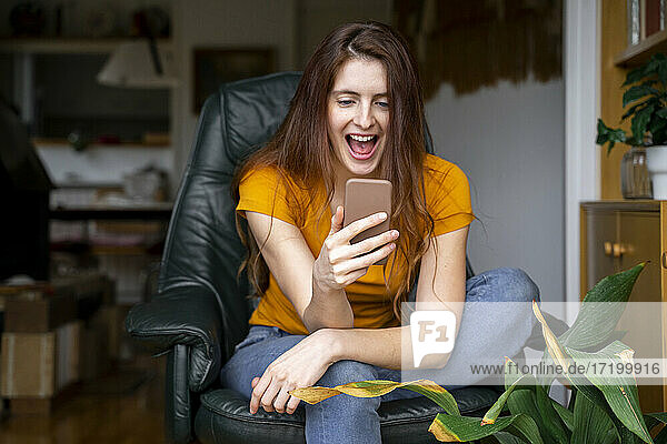 Fröhliche Frau  die ein Smartphone benutzt  während sie zu Hause auf einem Stuhl sitzt