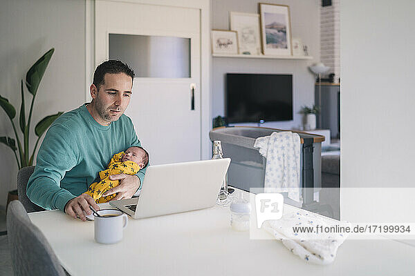Alleinstehender Vater arbeitet am Laptop  während er seinen Sohn am Schreibtisch im Heimbüro trägt