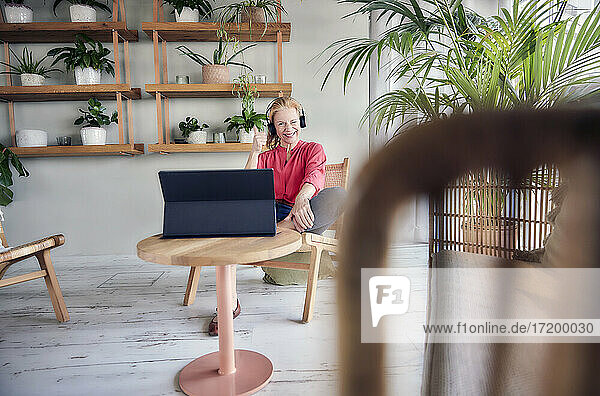 Lächelnde Frau  die zwinkert und Daumen nach oben zeigt  während sie ein Video auf einem Laptop in einer Wohnung ansieht