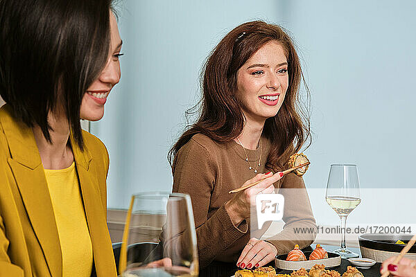 Lächelnde junge Frau isst Sushi  während sie mit Freundinnen an einem Tisch im Restaurant sitzt