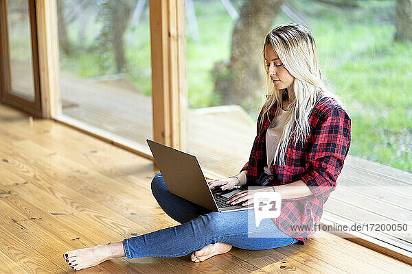 Blonde Frau arbeitet am Laptop und sitzt am Fenster im Vorgarten