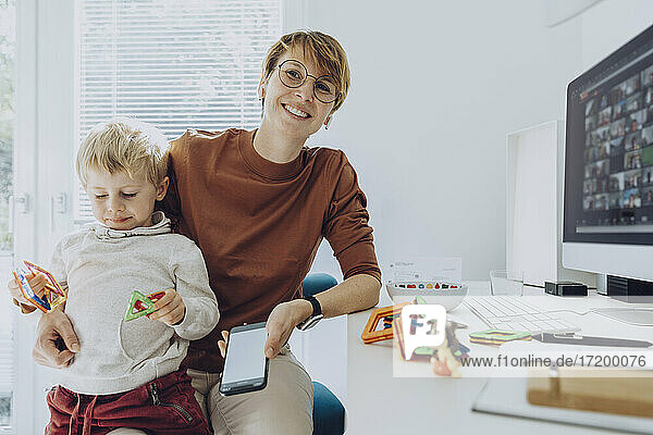 Smiling mother with son sitting on lap playing with toys in home office