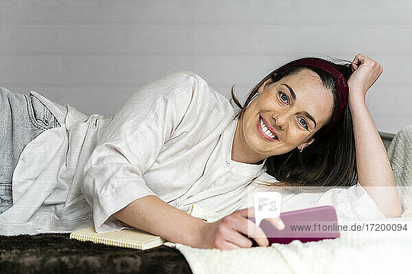 Schöne lächelnde Frau mit Smartphone auf dem Bett liegend