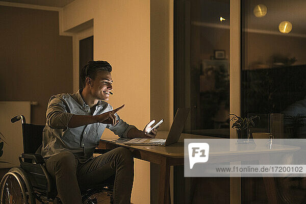 Smiling young man in wheelchair gesturing on video conference meeting while sitting on wheelchair in living room