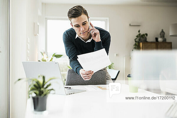 Young businessman holding document talking on smart while leaning on chair in home office