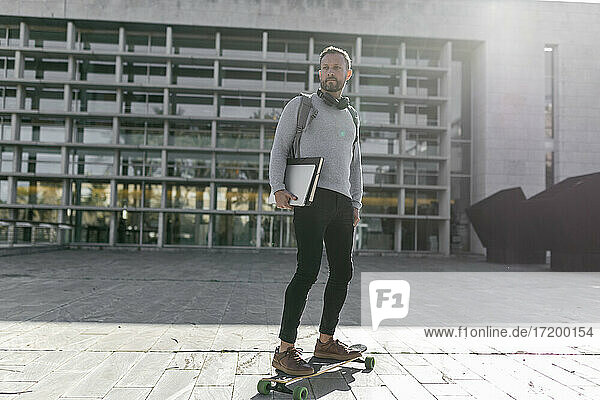 Männlicher Pendler mit Akten  der auf einem Skateboard vor einem Gebäude steht und wegschaut