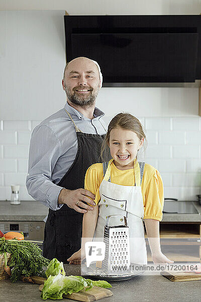 Vater und Tochter tragen eine Schürze und lächeln  während sie in der Küche zu Hause stehen