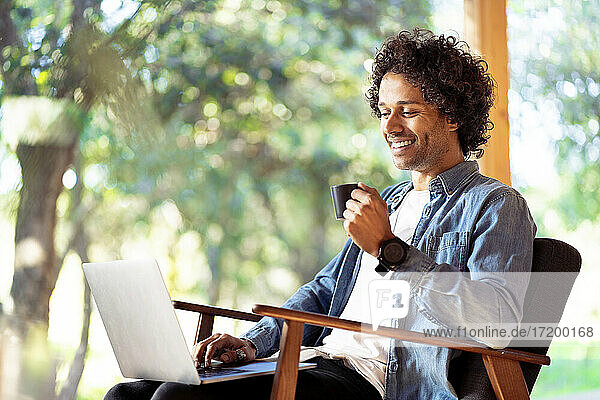 Smiling man with coffee cup using laptop while sitting on armchair at front yard