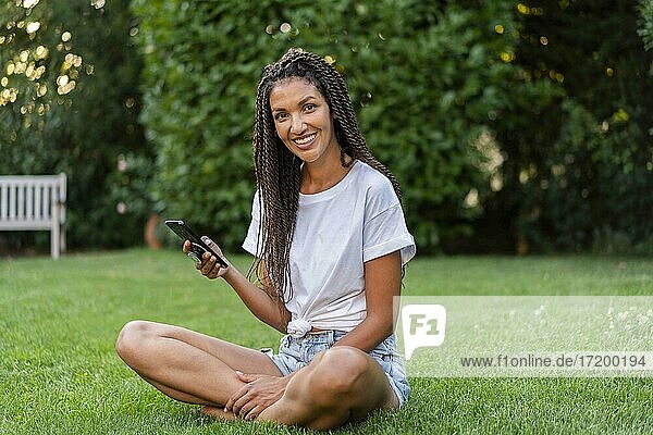 Lächelnde  geflochtene Frau mit Mobiltelefon im Schneidersitz im Garten
