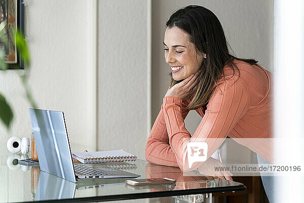 Happy female entrepreneur with head in hand leaning on dining table during video call at home