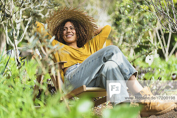 Lächelnde Frau mit Strohhut und Händen hinter dem Kopf entspannt sich auf einem Liegestuhl im Garten