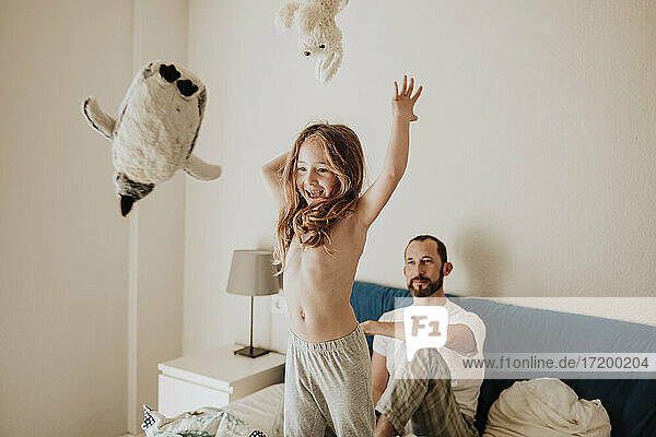 Fröhliches Mädchen wirft Plüschtiere  während der Vater zu Hause auf dem Bett zusieht