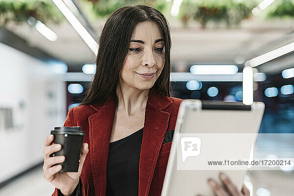 Lächelnde reife Geschäftsfrau mit Kaffeetasse  die auf ein digitales Tablet in einer U-Bahn-Station schaut