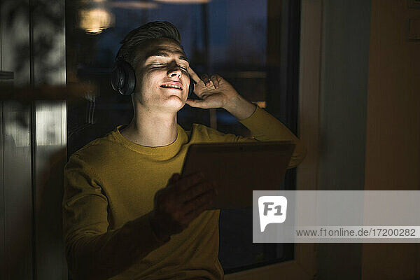 Junger Mann mit digitalem Tablet  der über drahtlose Kopfhörer Musik hört  während er im Wohnzimmer sitzt