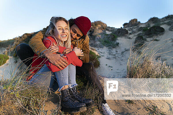 Affectionate couple smiling while sitting on sand dune during sunset