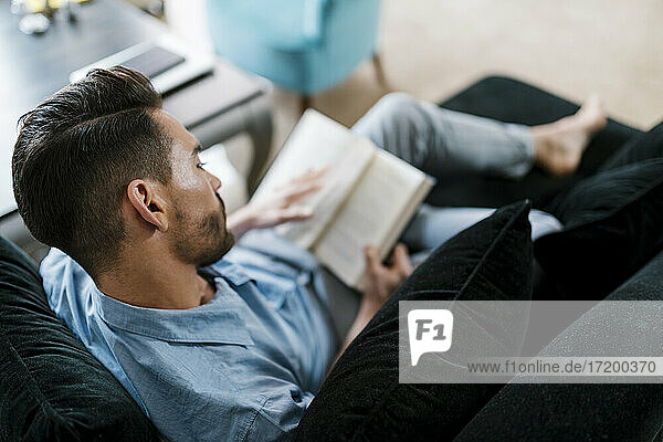 Mann liest ein Buch  während er zu Hause auf dem Sofa sitzt