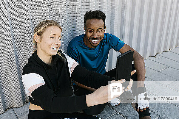 Fröhlicher männlicher und weiblicher Sportler  der ein Selfie durch ein digitales Tablet macht  während er auf dem Boden an der Wand sitzt