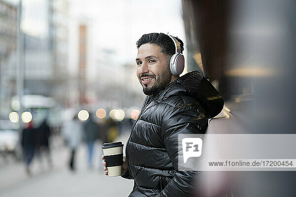 Lächelnder Hipster-Mann mit drahtlosen Kopfhörern  der eine Kaffeetasse hält und wegschaut