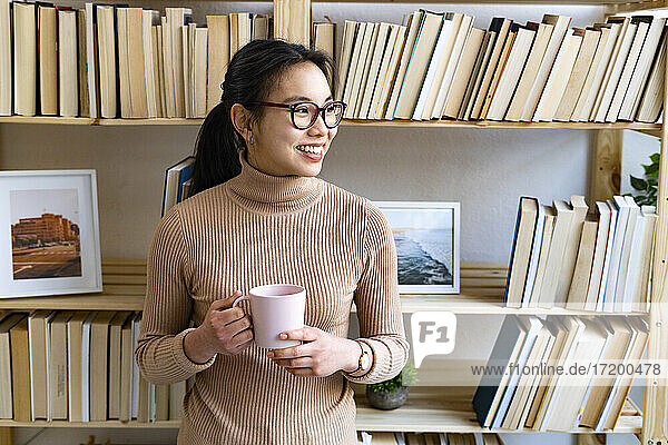 Woman with coffee cup smiling while standing by bookshelf at home