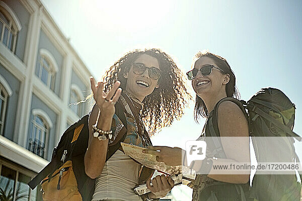 Cheerful female friends discussing over map while gesturing against sky on sunny day