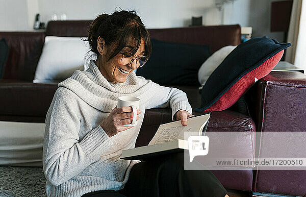 Frau mit Kaffeetasse lächelnd beim Lesen eines Buches zu Hause