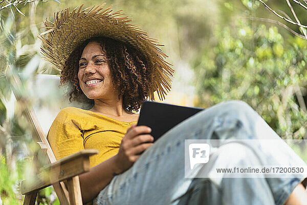 Lächelnde Frau mit Strohhut  die ein digitales Tablet hält und auf einem Stuhl im Gemüsegarten sitzt