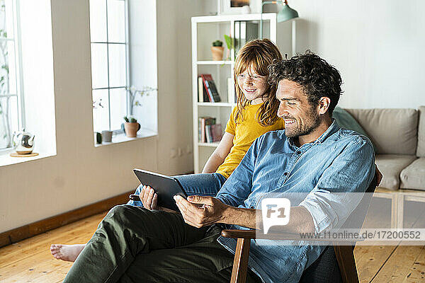 Vater und Tochter schauen auf ein digitales Tablet  während sie zu Hause auf einem Sessel sitzen