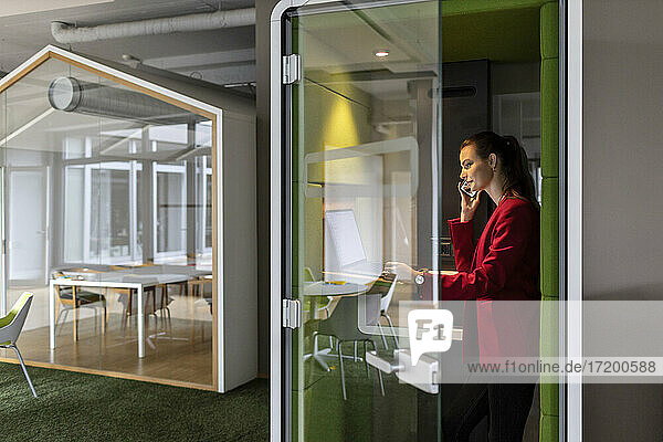 Lächelnde Unternehmerin  die einen Laptop benutzt  während sie in einer schalldichten Kabine im Büro mit einem Mobiltelefon spricht