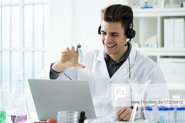 Lächelnde junge Wissenschaftlerin  die ein Reagenzglas während eines Videogesprächs auf einem Laptop im Labor zeigt