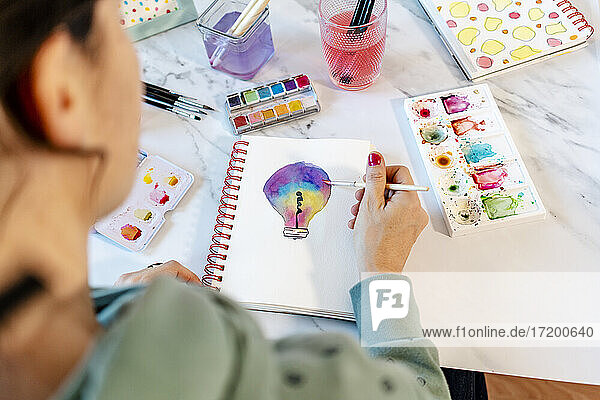 Woman painting light bulb with watercolors on spiral notebook