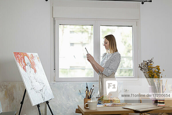 Weibliche Künstlerin  die ein Foto von einem Gemälde mit ihrem Smartphone macht  während sie im Heimstudio steht
