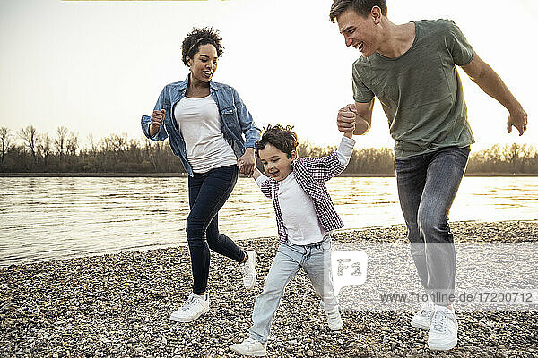 Junge hält die Hände seiner Eltern und läuft über einen Kieselstein am See