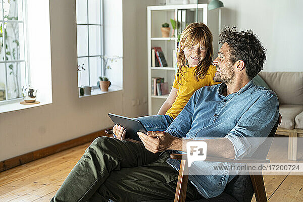 Vater mit digitalem Tablet  der seine Tochter betrachtet  die zu Hause auf einem Sessel sitzt