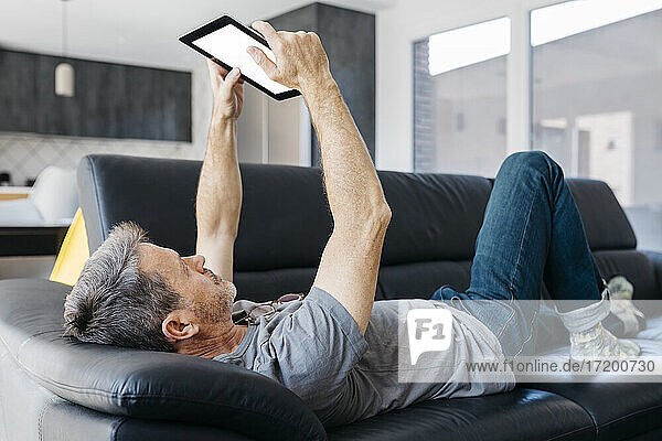 Mature man using digital tablet while lying on sofa at living room