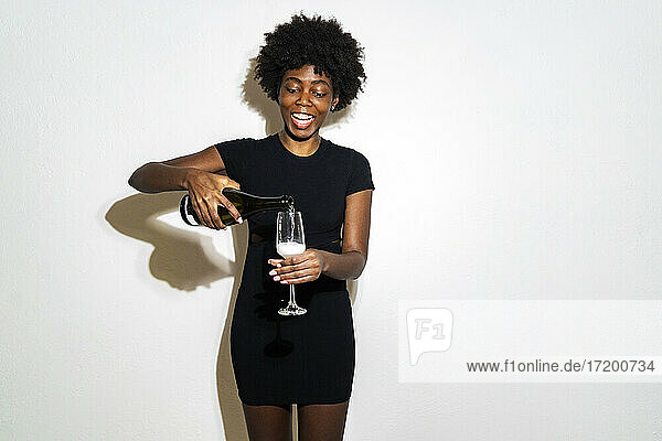 Glückliche junge Frau gießt Champagner in eine Champagnerflöte und steht vor einem weißen Hintergrund