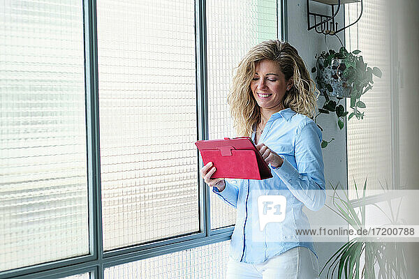 Lächelnde Frau  die ein digitales Tablet benutzt und sich gegen ein Glasfenster lehnt