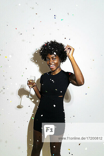 Confetti falling on happy woman dancing with champagne flute while standing against white background