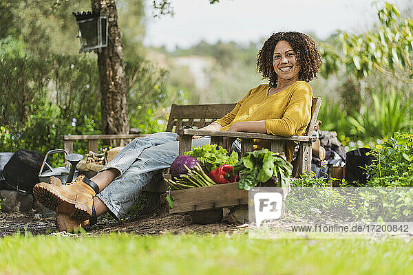 Junge lächelnde Frau sitzt auf einer Holzbank in einem nachhaltigen Permakultur-Garten