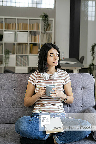 Nachdenkliche junge Frau mit Kaffeetasse auf dem Sofa sitzend und wegschauend