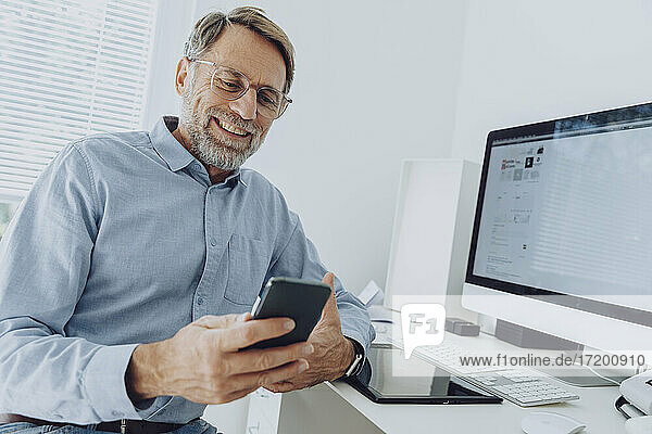 Lächelnder reifer Geschäftsmann mit Brille  der im Büro zu Hause ein Mobiltelefon benutzt