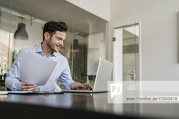Smiling male entrepreneur with document working on laptop in office
