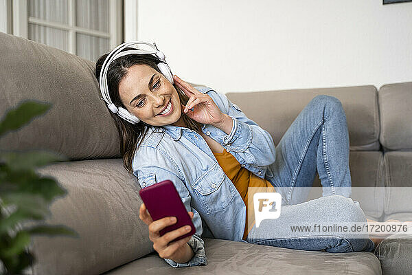 Lächelnde Frau  die ihr Handy in der Hand hält  während sie auf dem Sofa zu Hause über Kopfhörer Musik hört